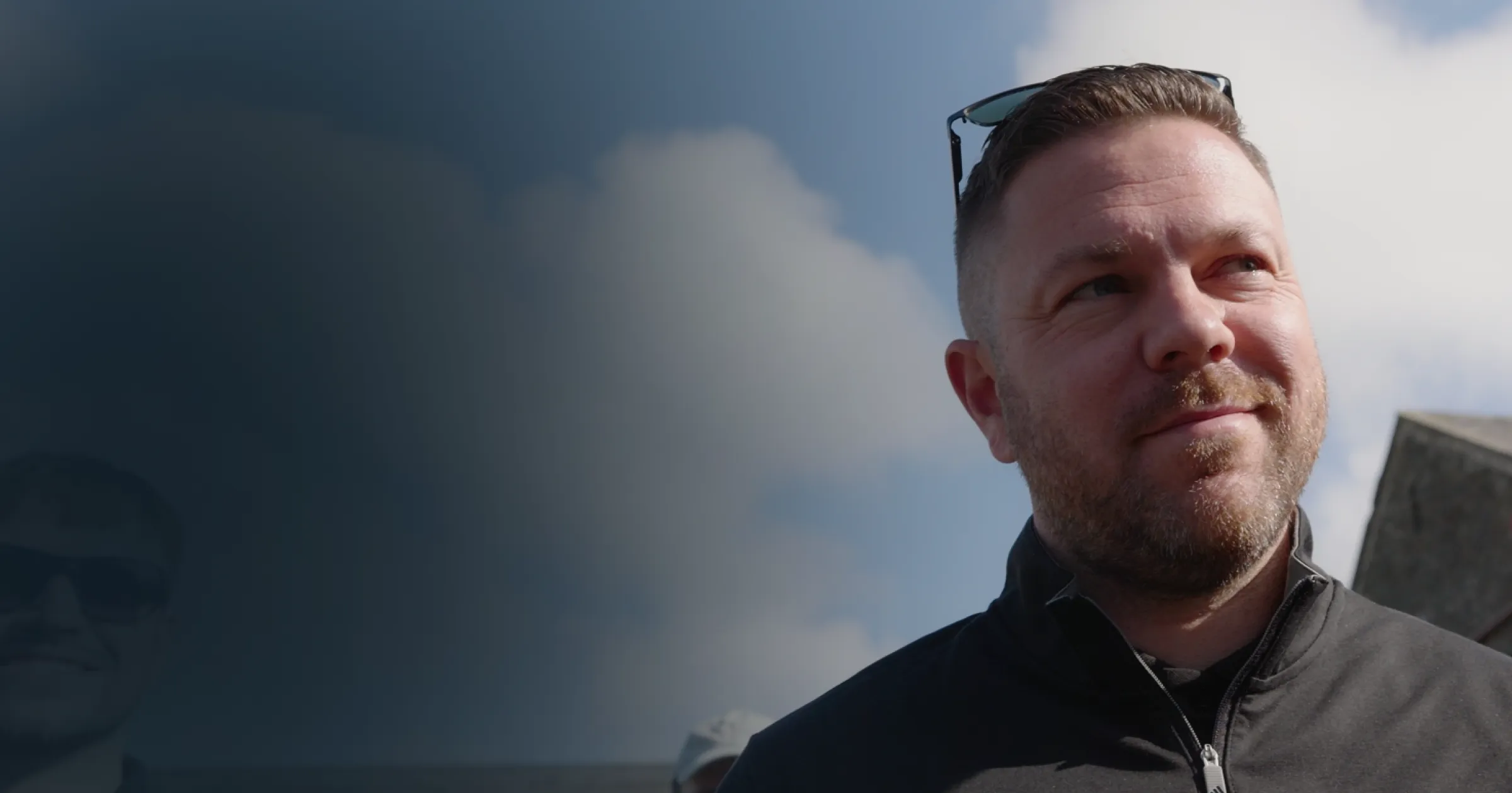 A man with short hair and a beard stands under a partly cloudy sky, smiling slightly. He wears sunglasses on his head and a zipped-up jacket. The mood is reflective.